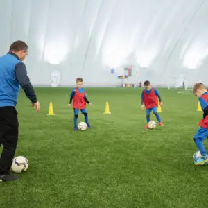 Coach training kids in soccer drills indoors on artificial turf.
